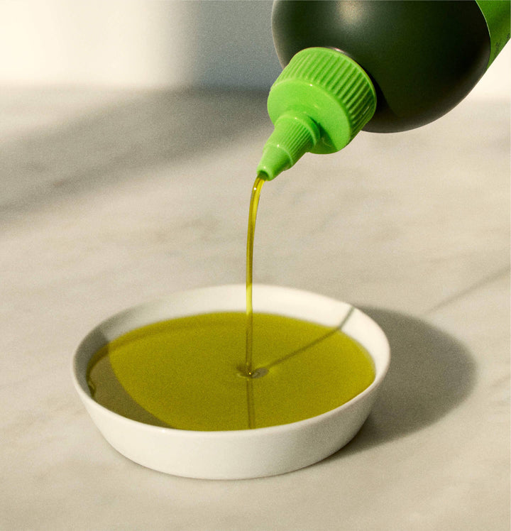 Green bottle pouring oil into a white bowl on a light surface