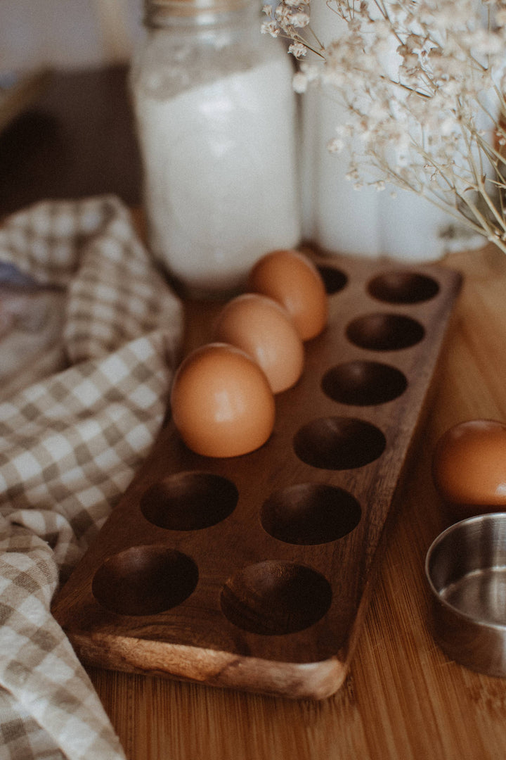 Wooden egg holder with eggs on a wooden surface with a checkered cloth and jars in the background.
