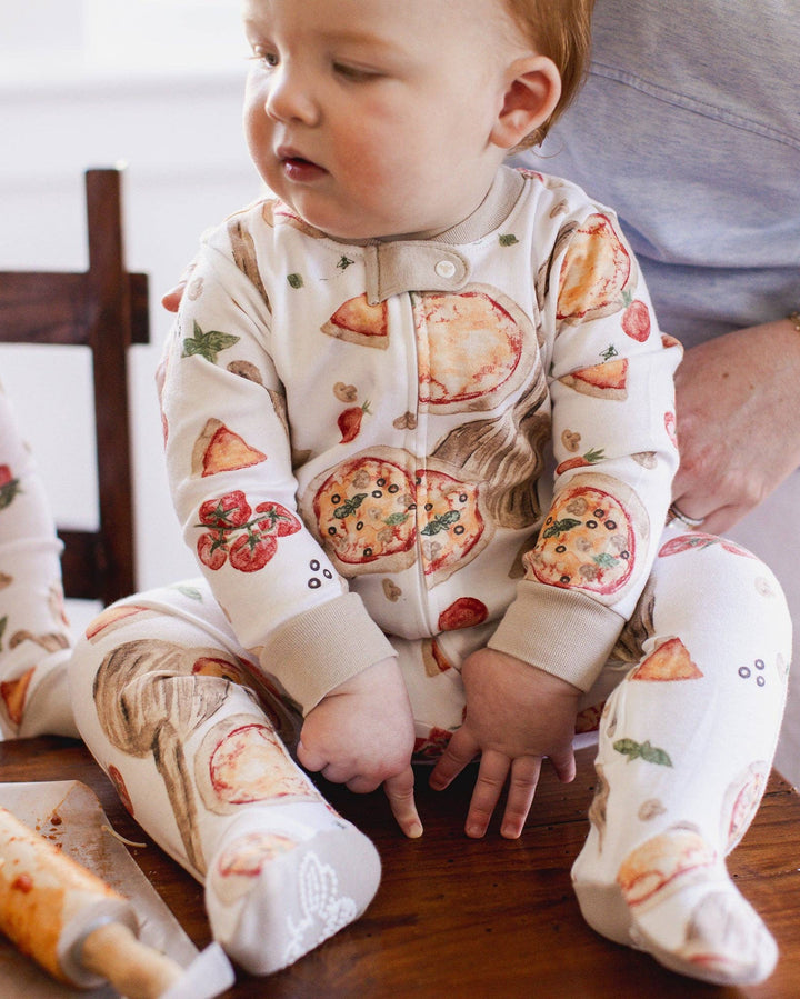 Baby wearing a pizza-themed onesie sitting at a table with pizza ingredients.