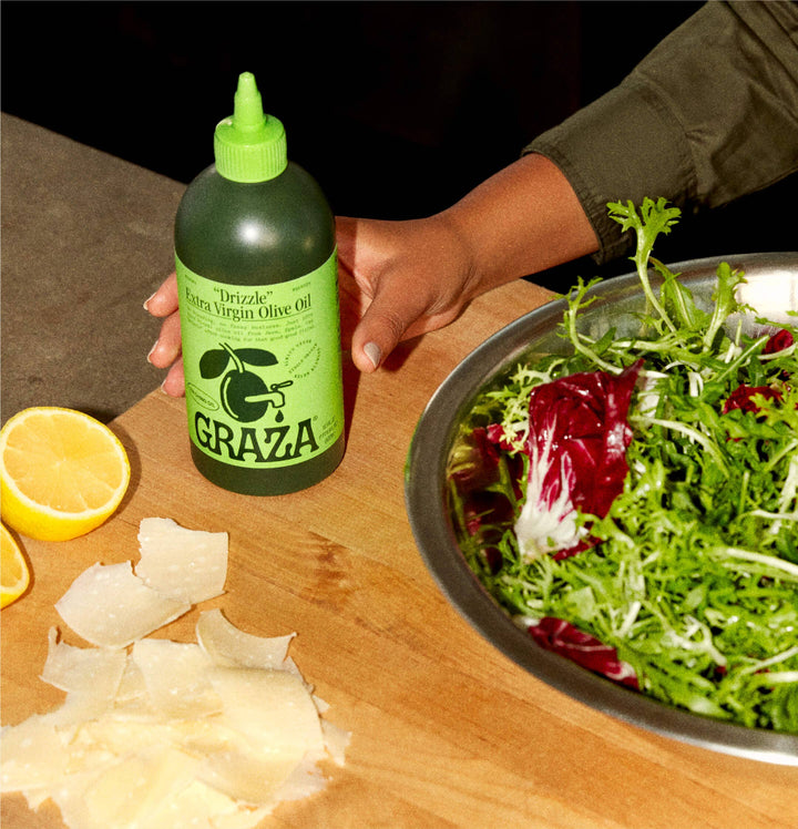 Person holding a bottle of Graza olive oil next to a salad on a wooden table.