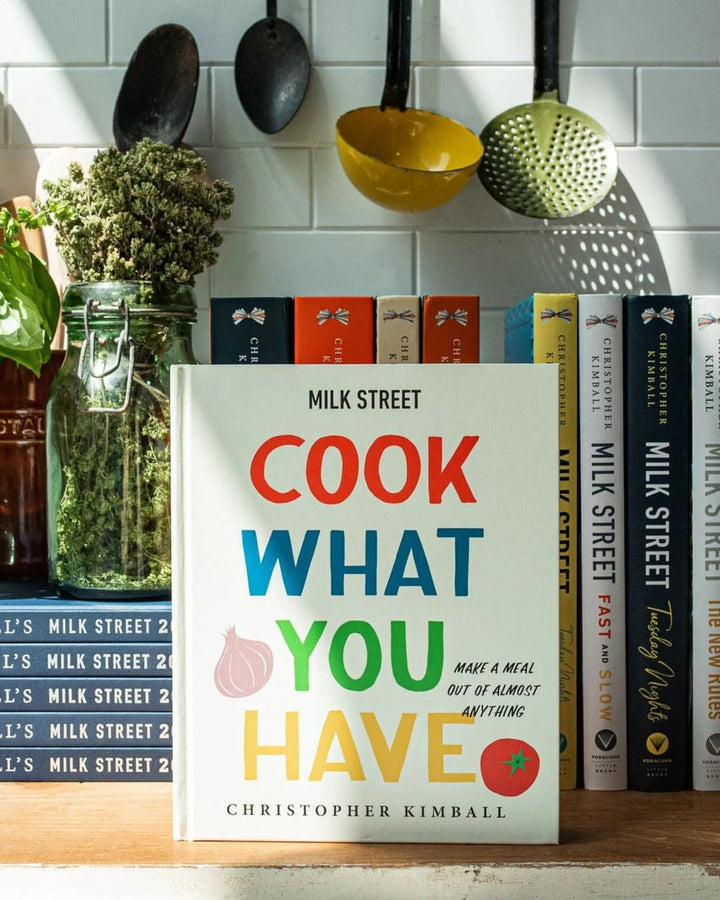 Cookbook titled 'Cook What You Have' by Christopher Kimball on a kitchen shelf with cookbooks and kitchen items.