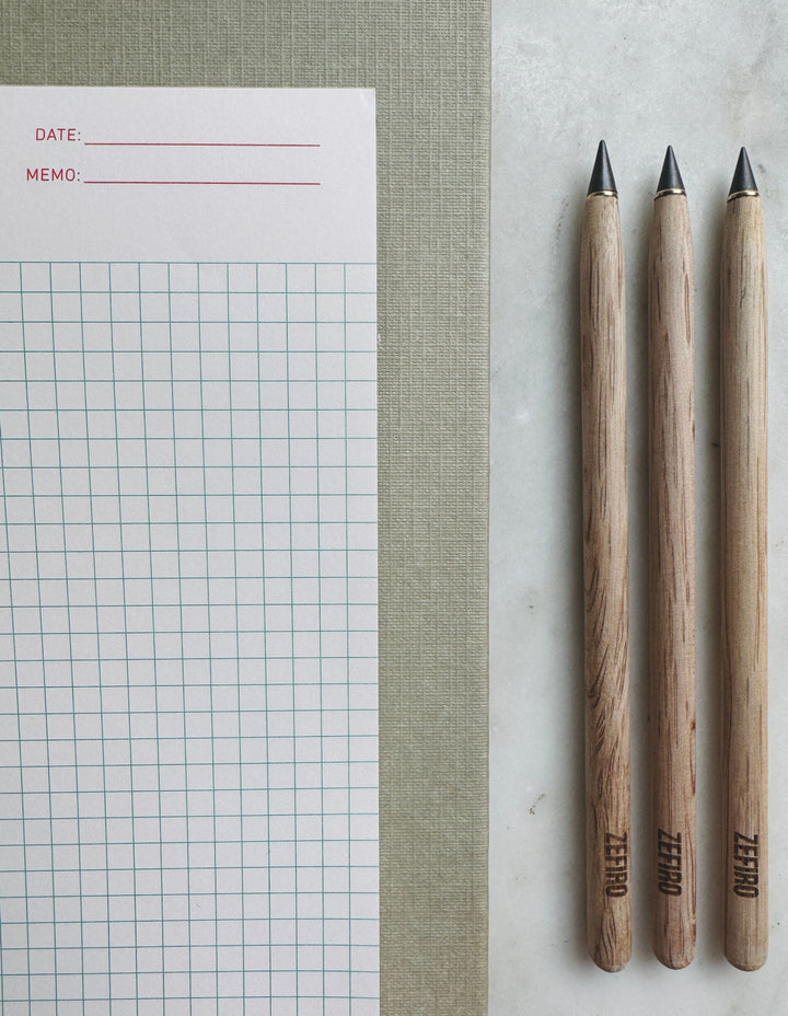 Three wooden pencils with branded logo next to a piece of paper with grid and memo lines on a textured surface.