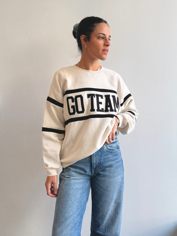 Woman wearing a cream sweatshirt with 'GO TEAM' text and blue jeans against a plain background