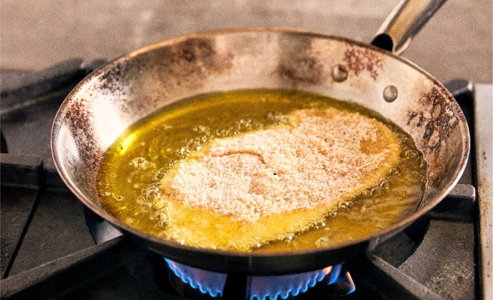 Breaded chicken bubbling in a frying pan filled with olive oil on a gas stove