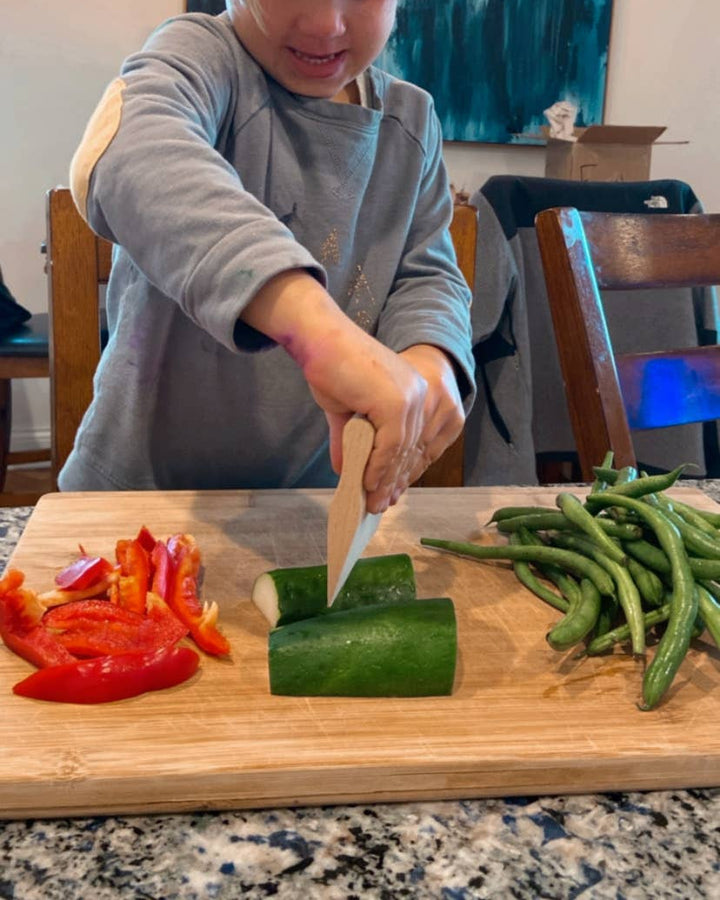Child cutting vegetables using a wooden paddle knifen on a wooden cutting board in a kitchen.