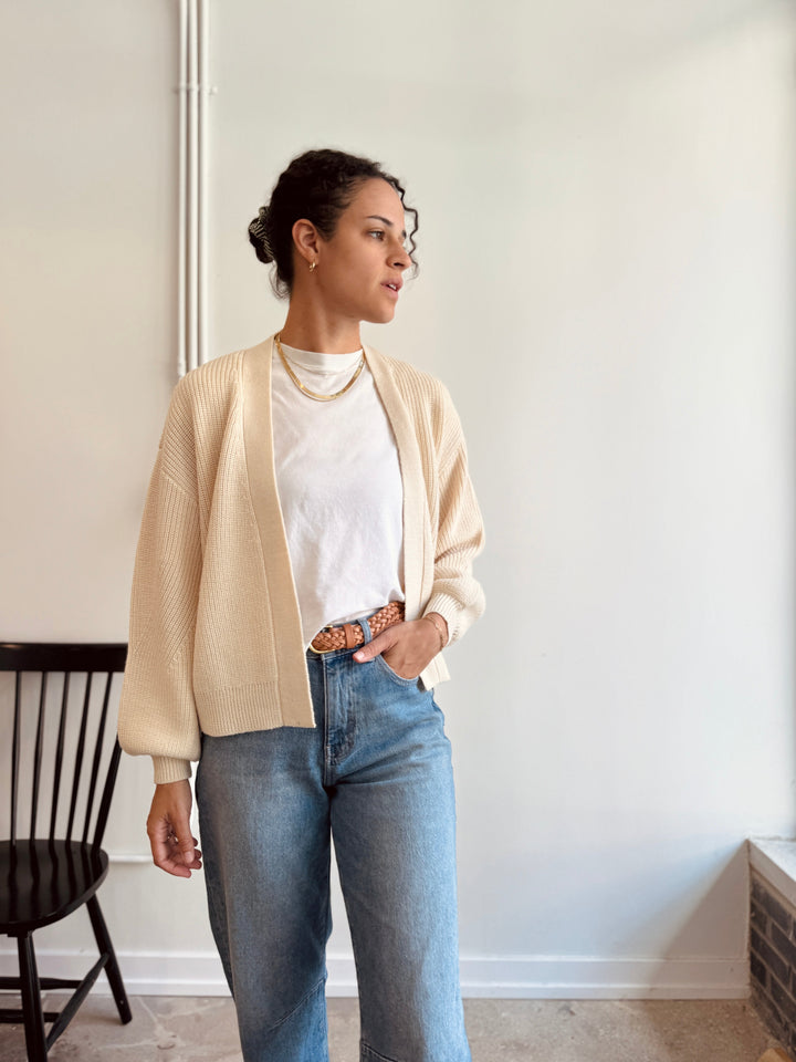 Woman wearing a beige cardigan, white shirt, and blue jeans indoors.