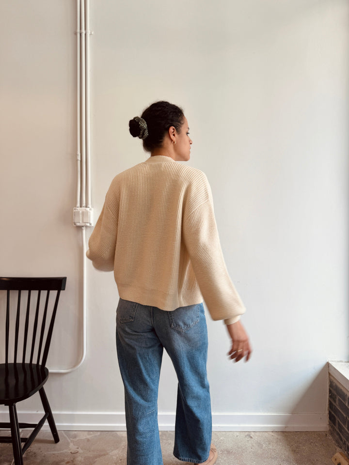 Woman wearing a beige sweater and blue jeans standing against a white wall.