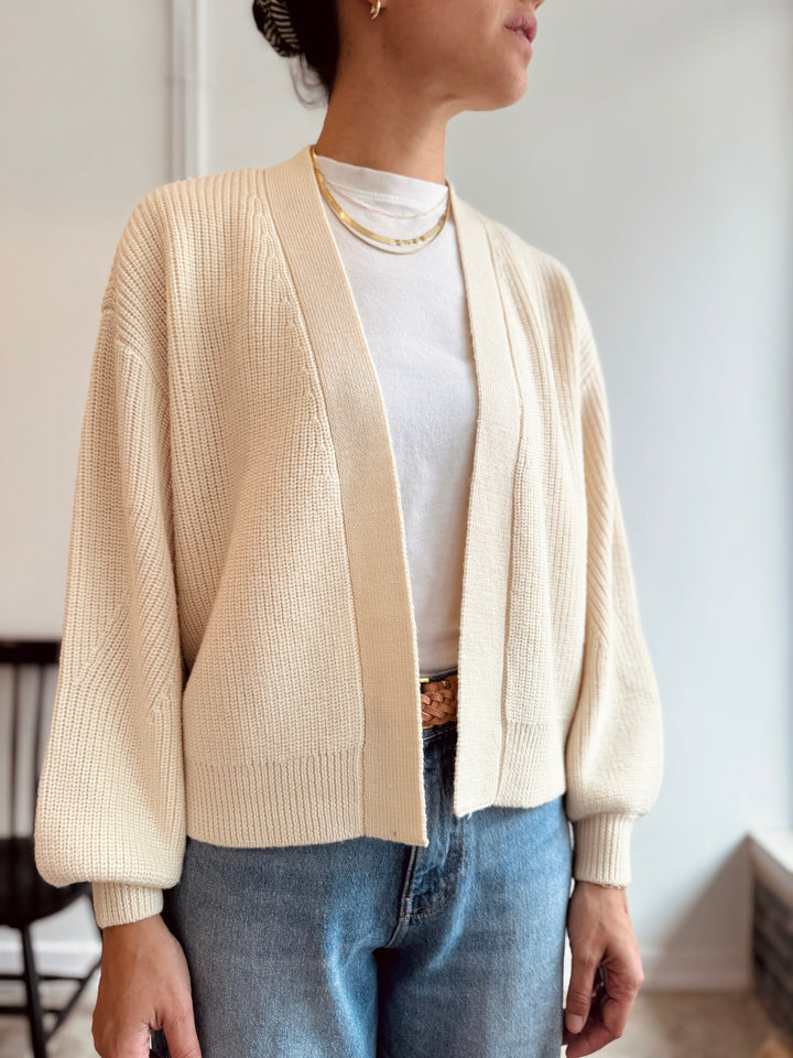 Person wearing a beige cardigan over a white shirt with blue jeans indoors.