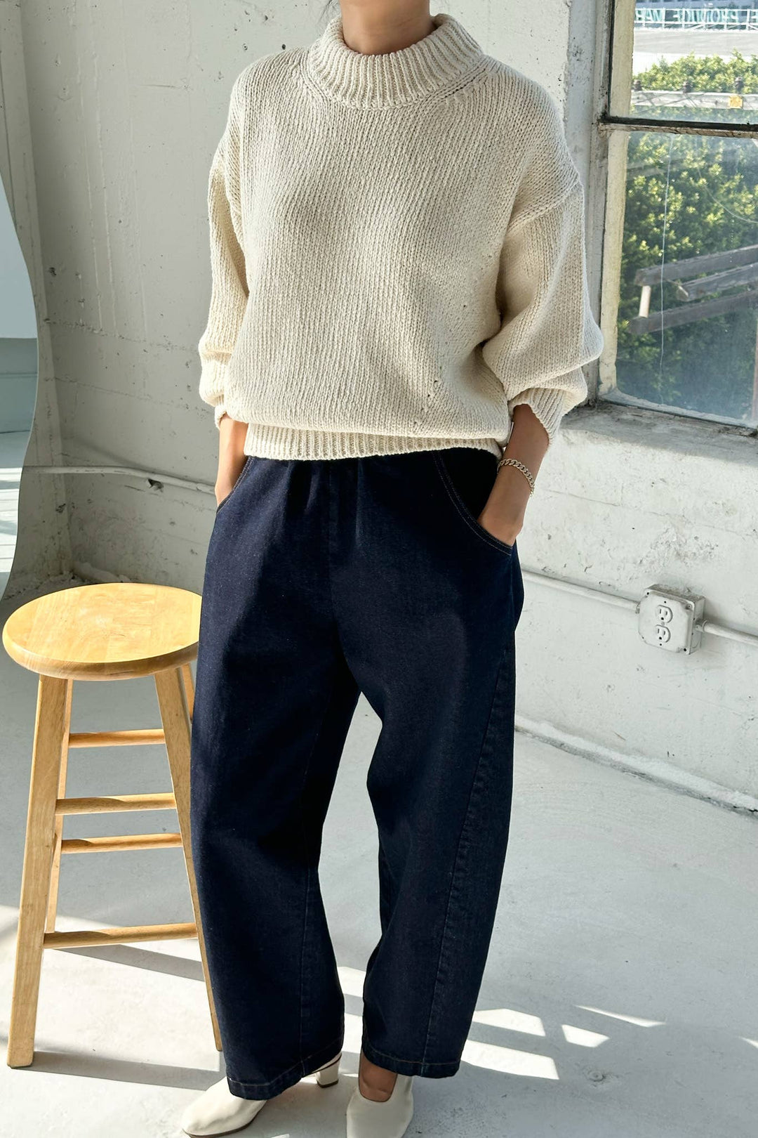Person wearing a beige sweater and dark blue pants standing indoors with a window in the background.