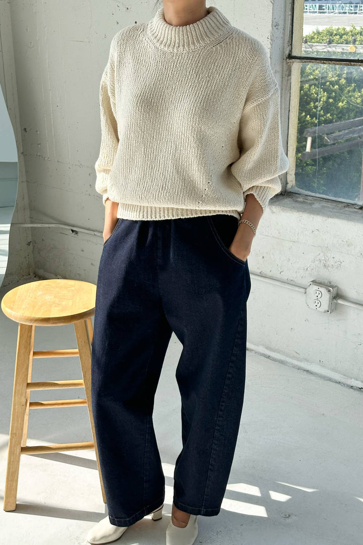 Person wearing a beige sweater and dark blue pants standing indoors with a window in the background.