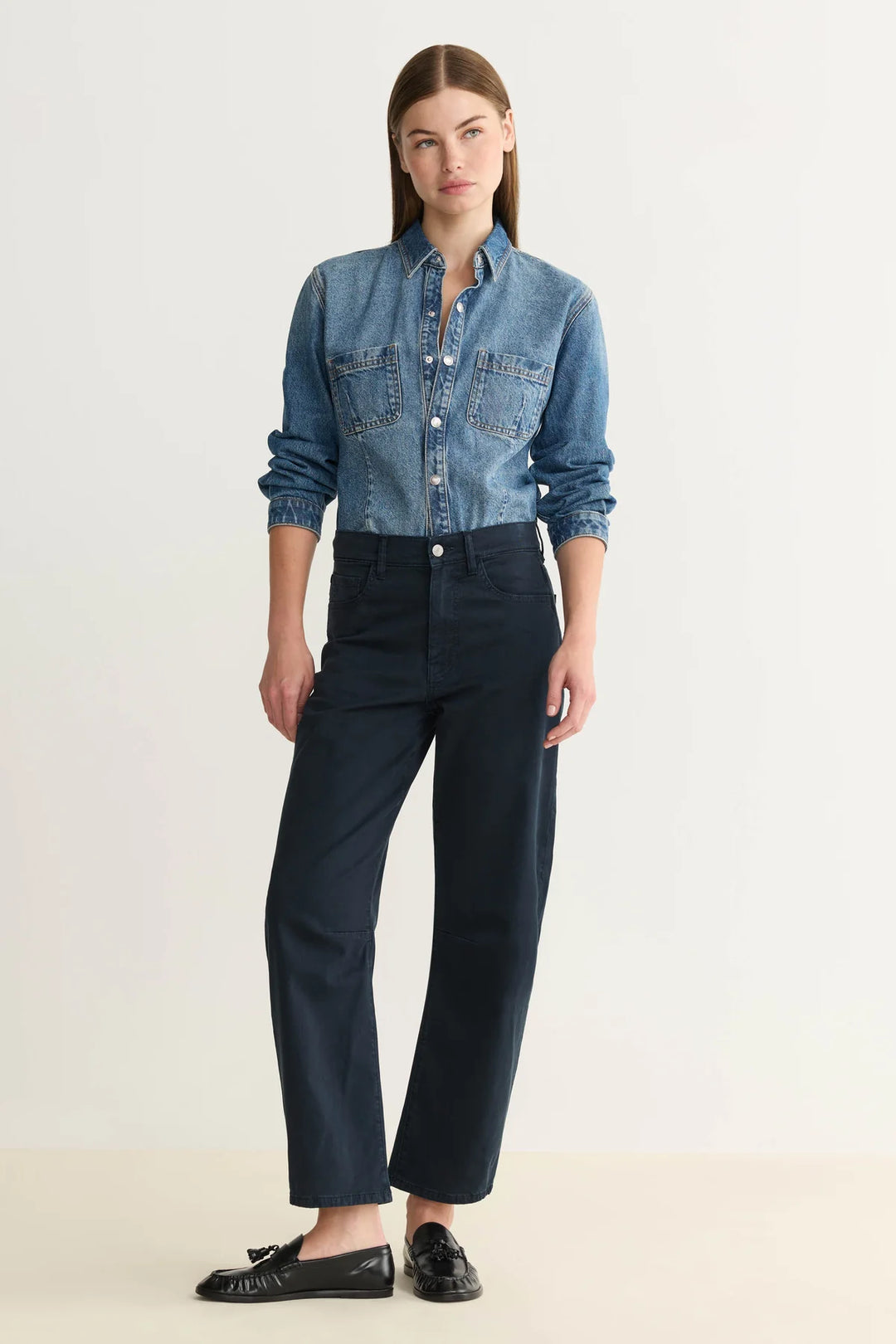 Woman wearing a blue denim shirt and navy blue barrel leg pants on a white background