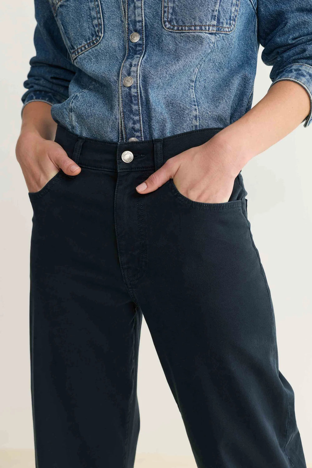 Person wearing a blue denim shirt tucked into navy blue pants on a plain background