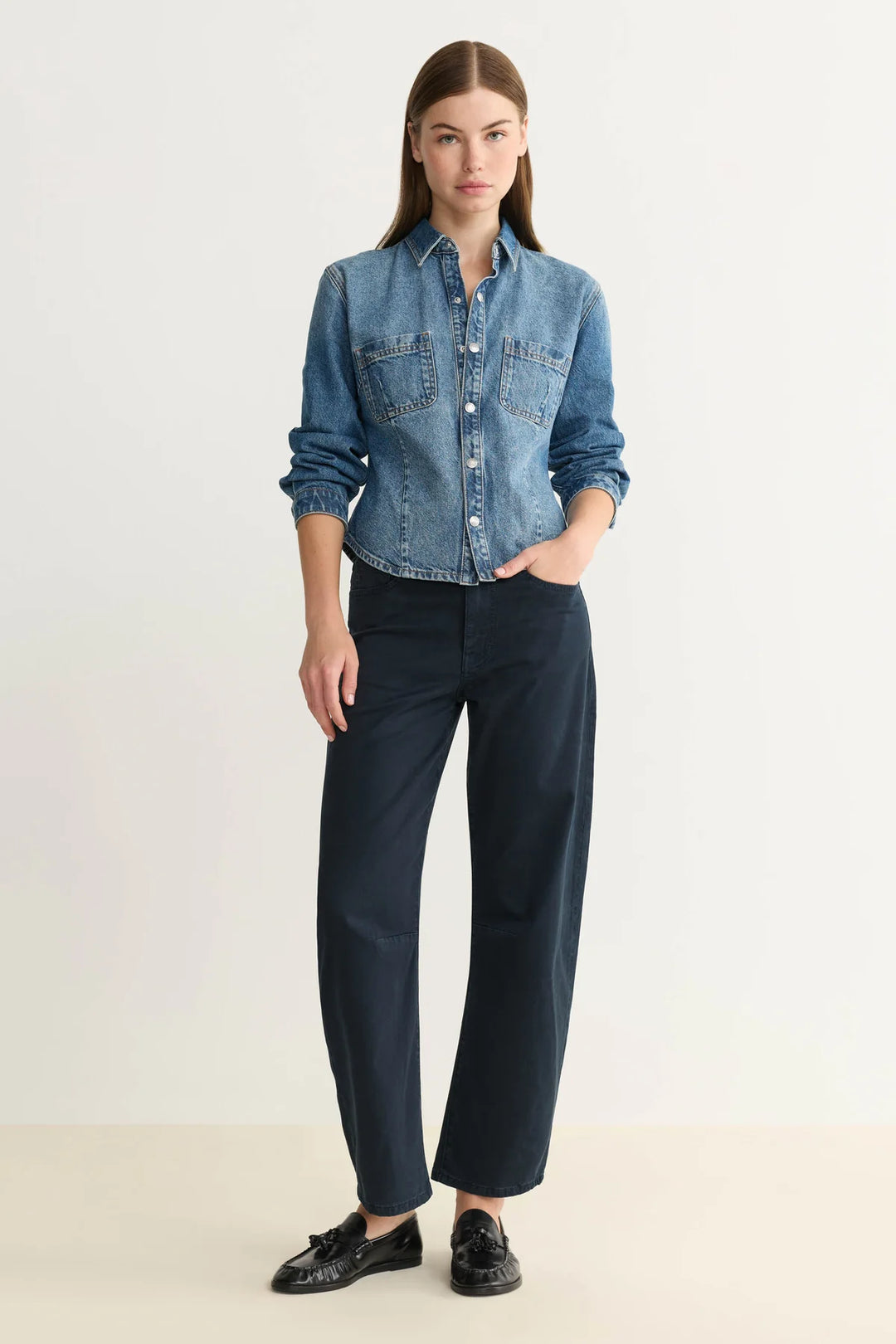 Woman wearing a blue denim shirt and navy blue barrel leg pants on a white background