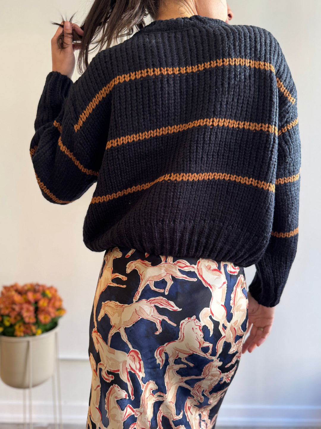 Person wearing a dark blue sweater with orange stripes and patterned pants, standing against a white wall.