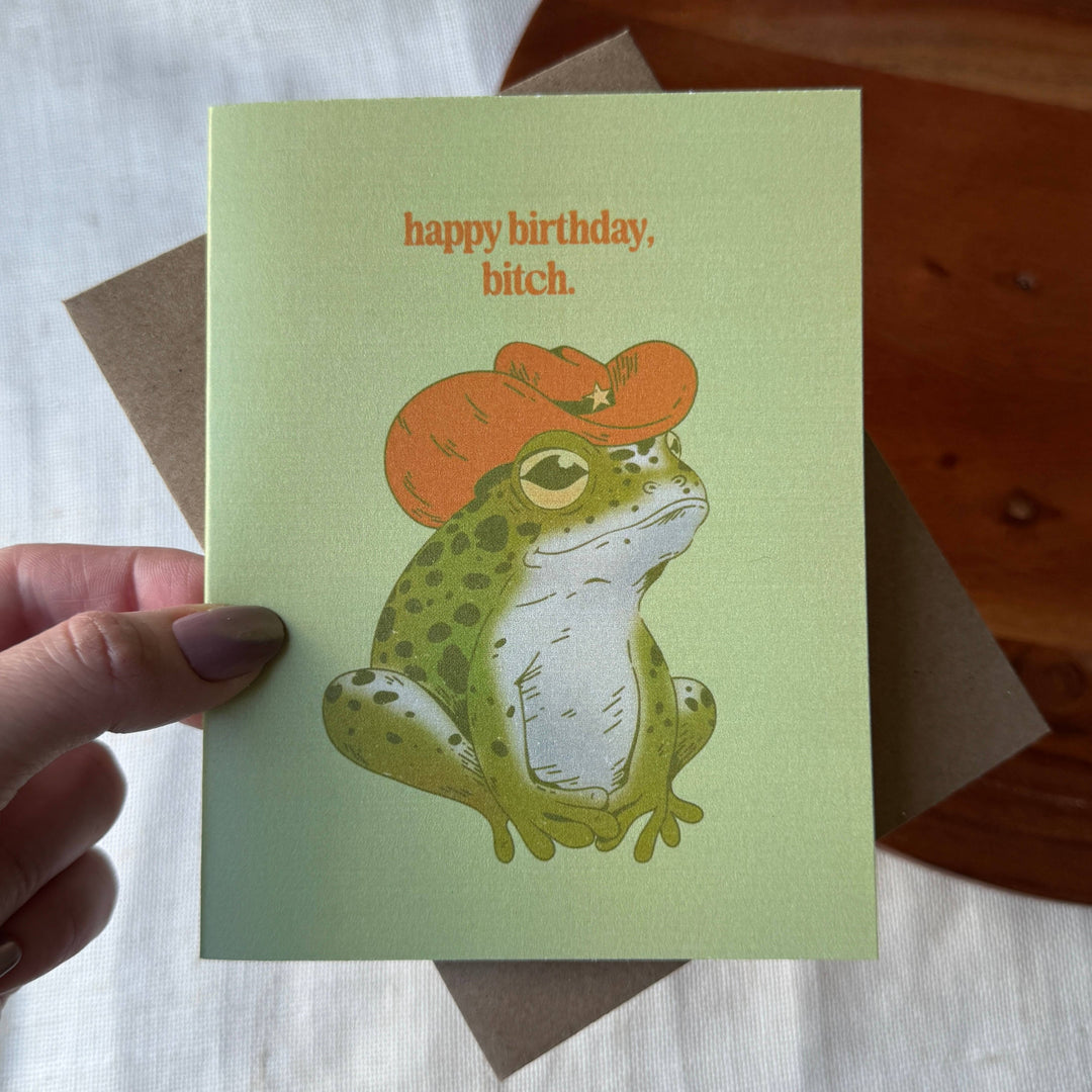 Card with a frog wearing a red hat and 'happy birthday, bitch.' text held by a hand.