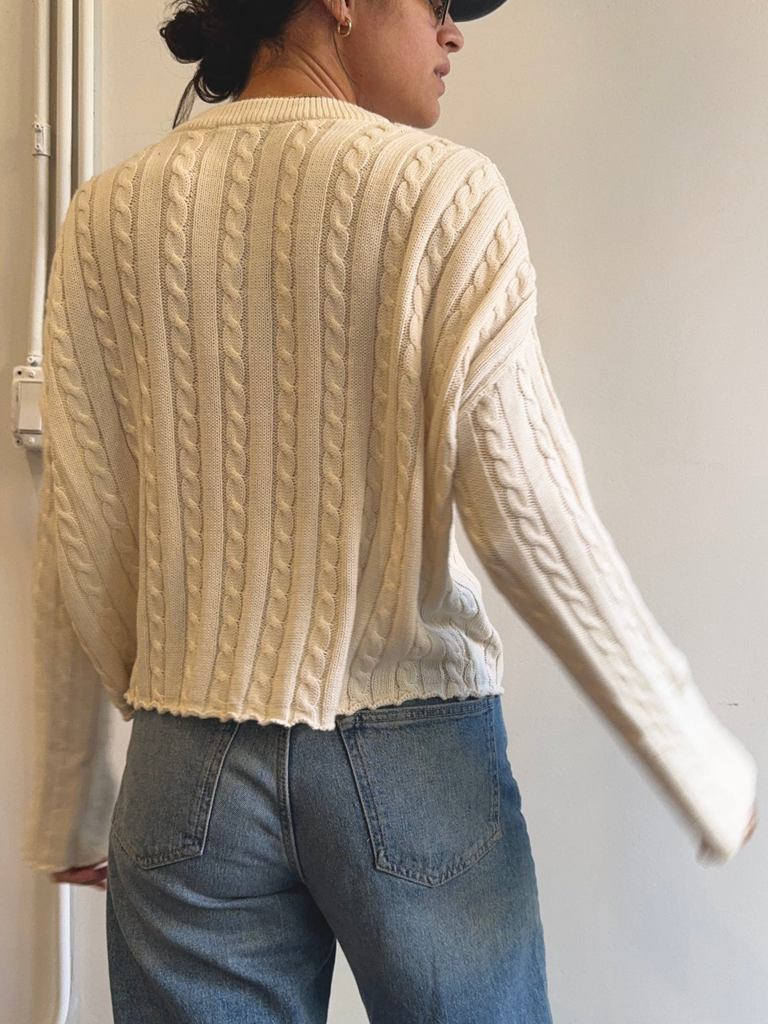 Person wearing a beige cable knit sweater and blue jeans against a plain background