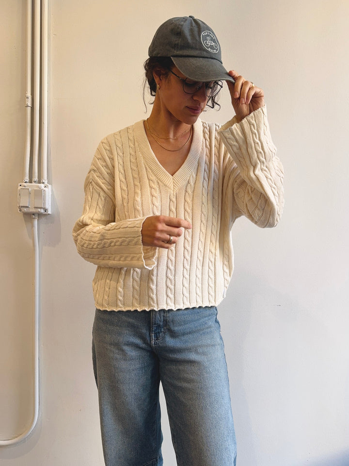 Person wearing a cream cable knit sweater and blue jeans, adjusting a dark cap against a plain background.