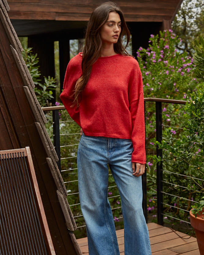 Woman wearing a red sweater and blue jeans standing on a wooden deck with greenery in the background