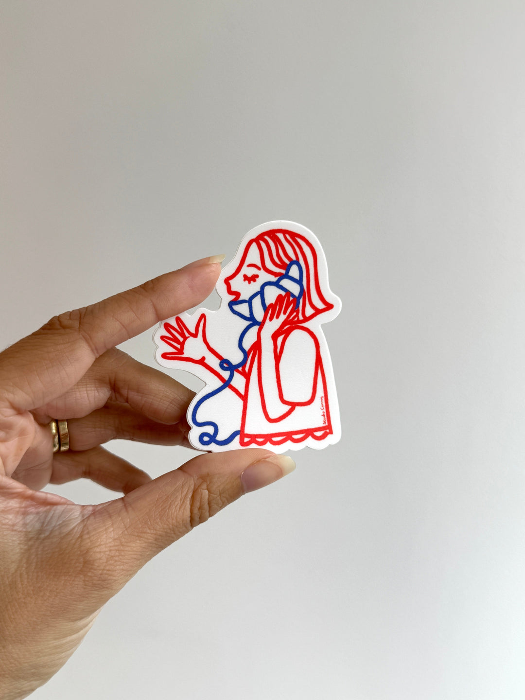 A hand holds a die-cut sticker featuring a minimalist line art illustration of a woman in profile, drawn with red and blue lines on a white sticker backing, against a plain light gray background.