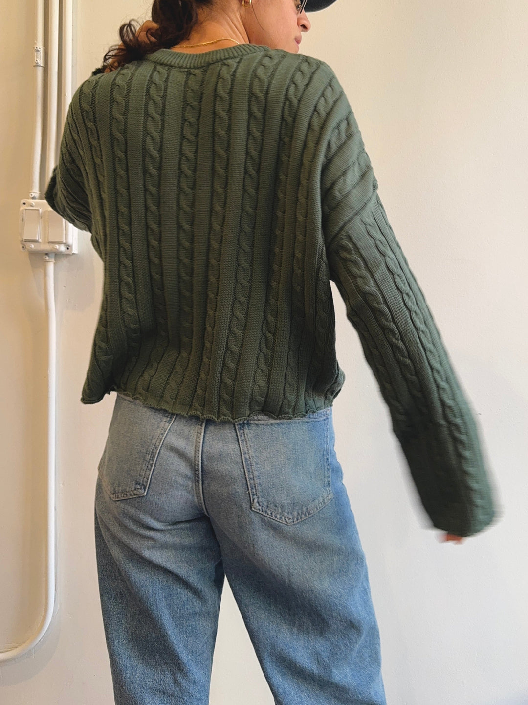 Person wearing a green cable knit sweater and blue jeans against a neutral background