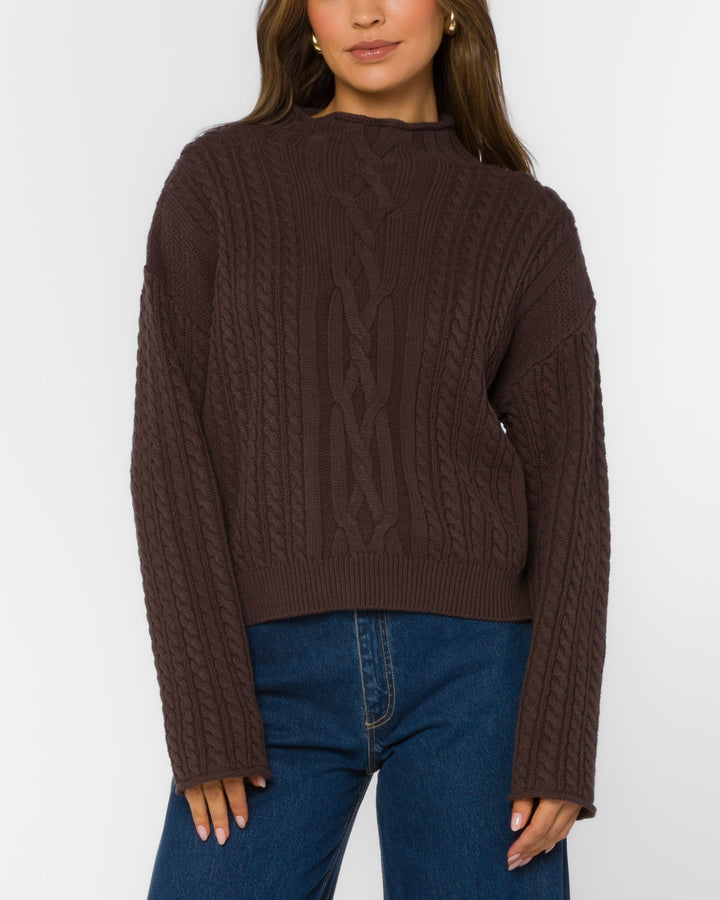 Woman wearing a brown cable knit sweater and blue jeans on a white background