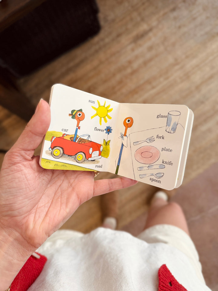 Hand holding a small open book with illustrations on a wooden floor.