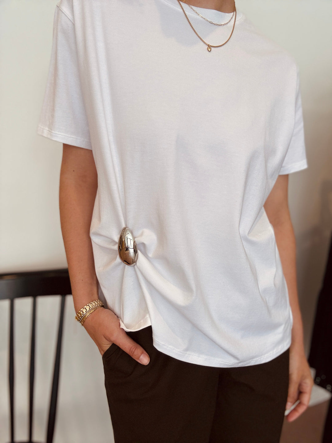 Person wearing a loose white tee with a gold drip shaped brooch pinning up the lower hem of the tee. Available at winsome clothing & goods in Avondale, Chicago.