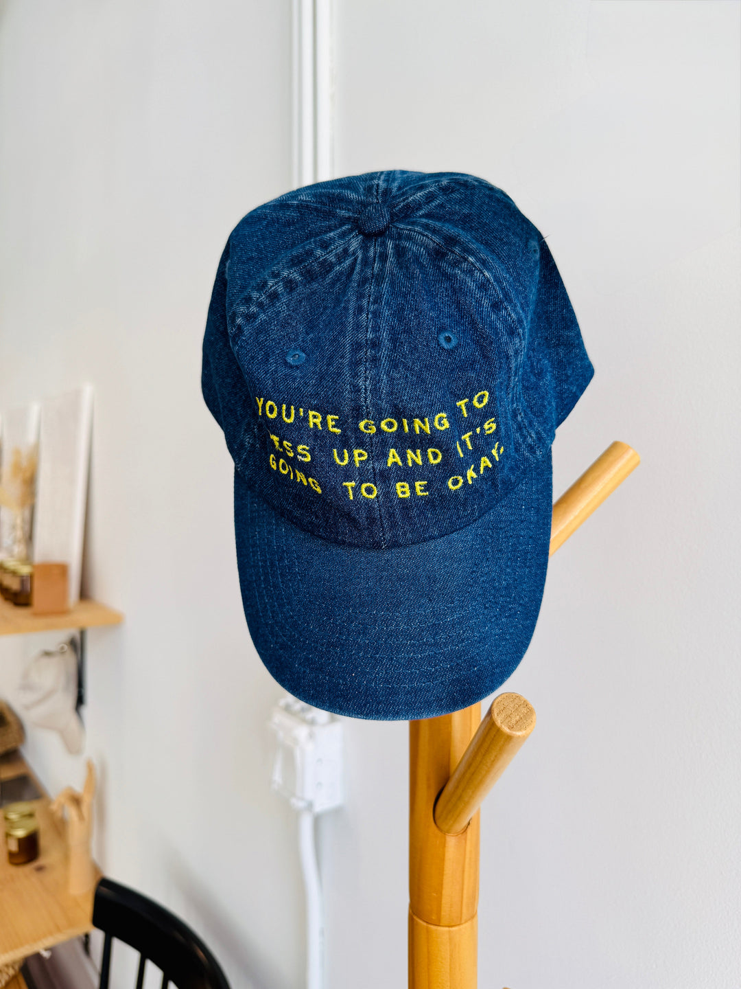 Blue denim cap with yellow text on a wooden rack against a white wall.
