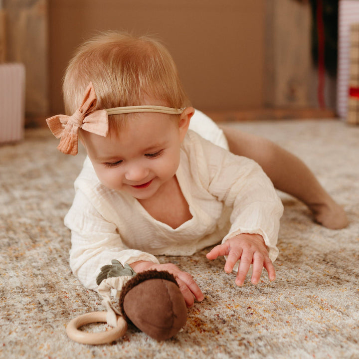 Baby crawling on a carpeted floor with an acorn toy, wearing a white outfit and headband.