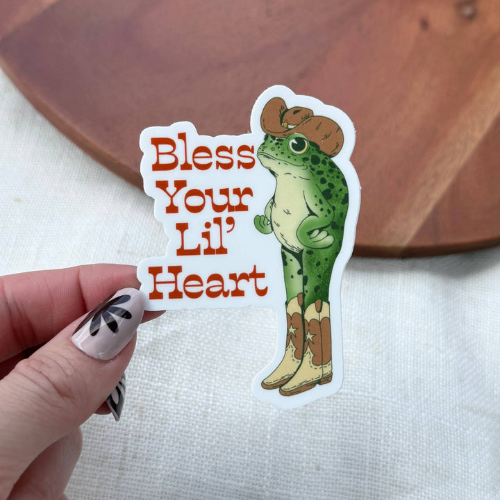 Sticker with a frog wearing boots and a cowboy hat, text 'Bless Your Lil' Heart', held by a hand.