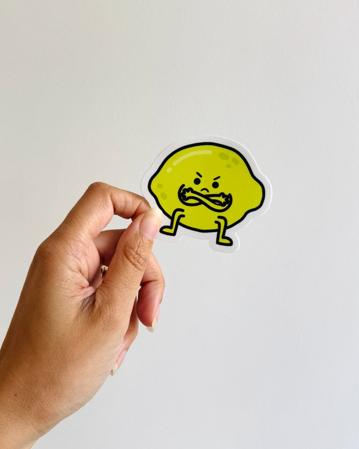A hand holding a die-cut sticker of a grumpy cartoon lemon against a plain light gray background.