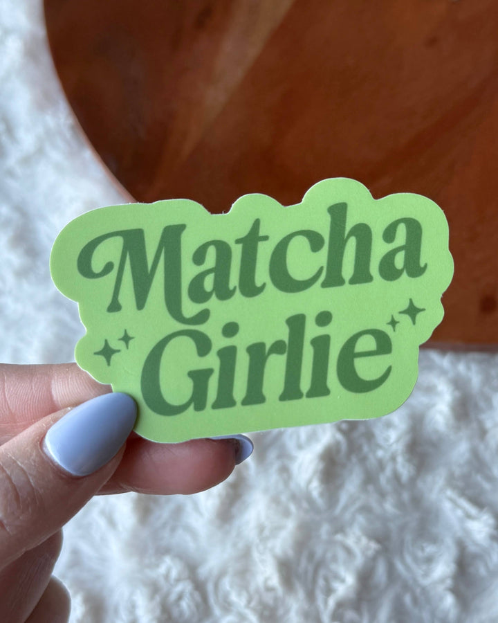 Hand holding a green 'Matcha Girlie' sticker against a textured white and wooden background