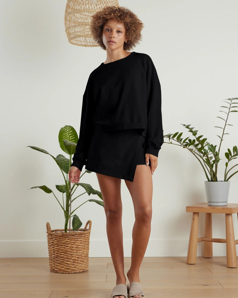 Person wearing a black outfit standing in a room with plants and a wooden stool.