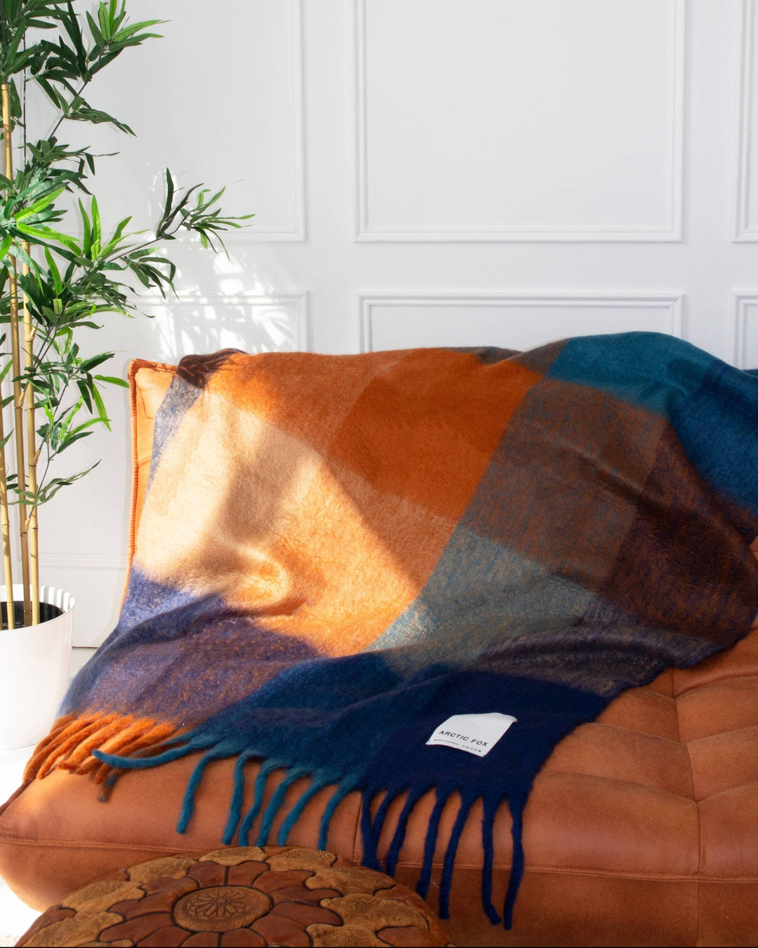 Colorful blanket draped over a brown leather chair with a white wall and plant in the background