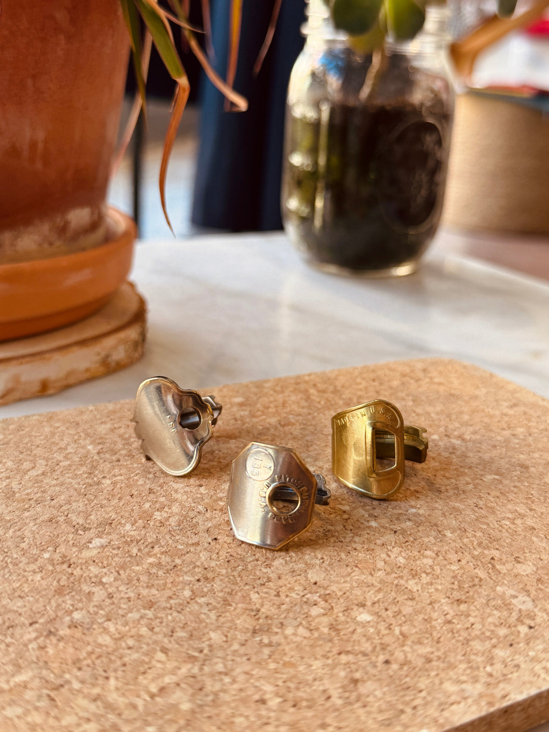 Three vintage keys that have been wrapped into rings in silver and gold, resting on a cork surface on a marble tabletop with a blurred potted plant and glass jar in the background. Rings and jewelry available at Winsome a boutique in Avondale, Chicago