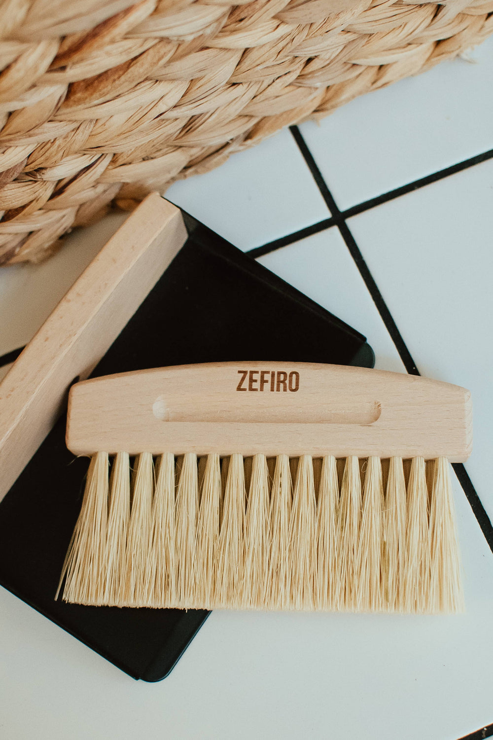 Wooden brush with bristles on a black surface, branded 'Zefiro', with a woven basket in the background.