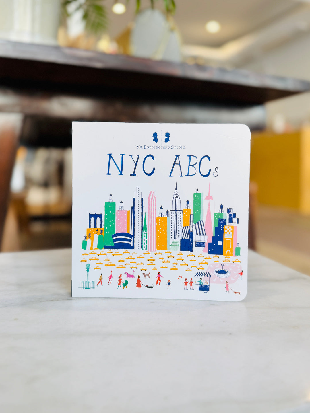 Children's book titled 'NYC ABCs' on a table with a blurred indoor background