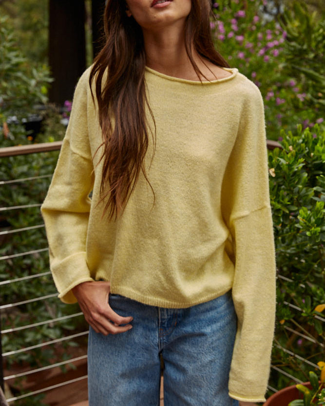 Woman wearing a yellow sweater and blue jeans standing outdoors with greenery in the background