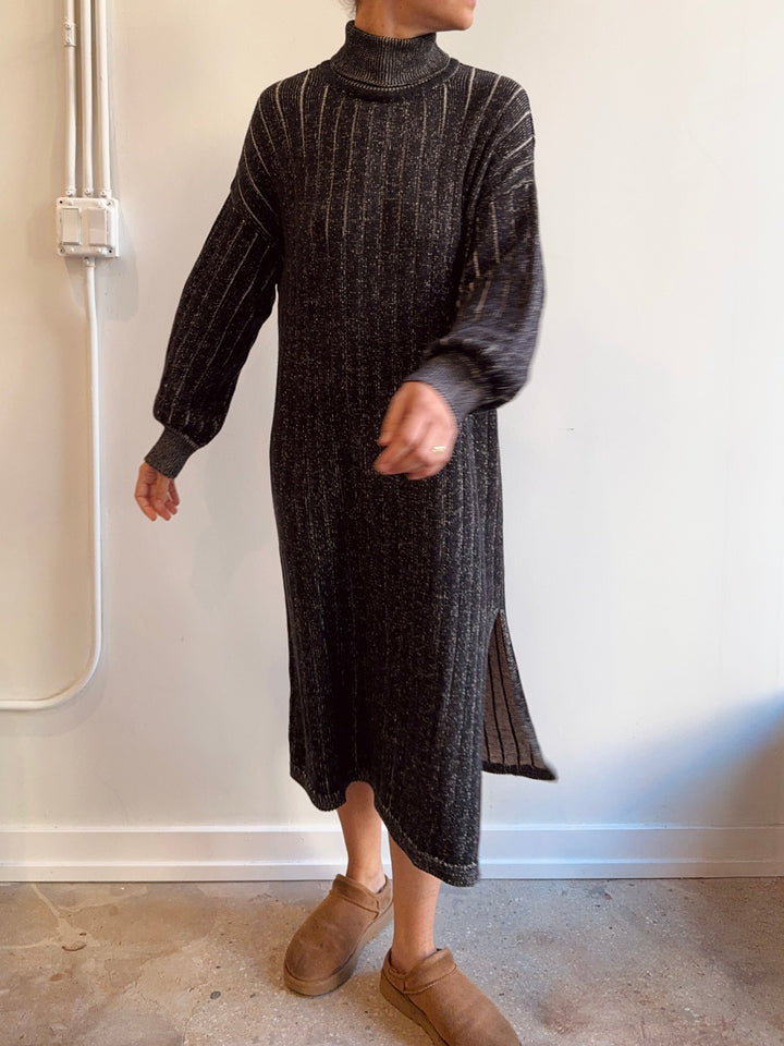 Person wearing a long dark ribbed sweater against a plain wall.