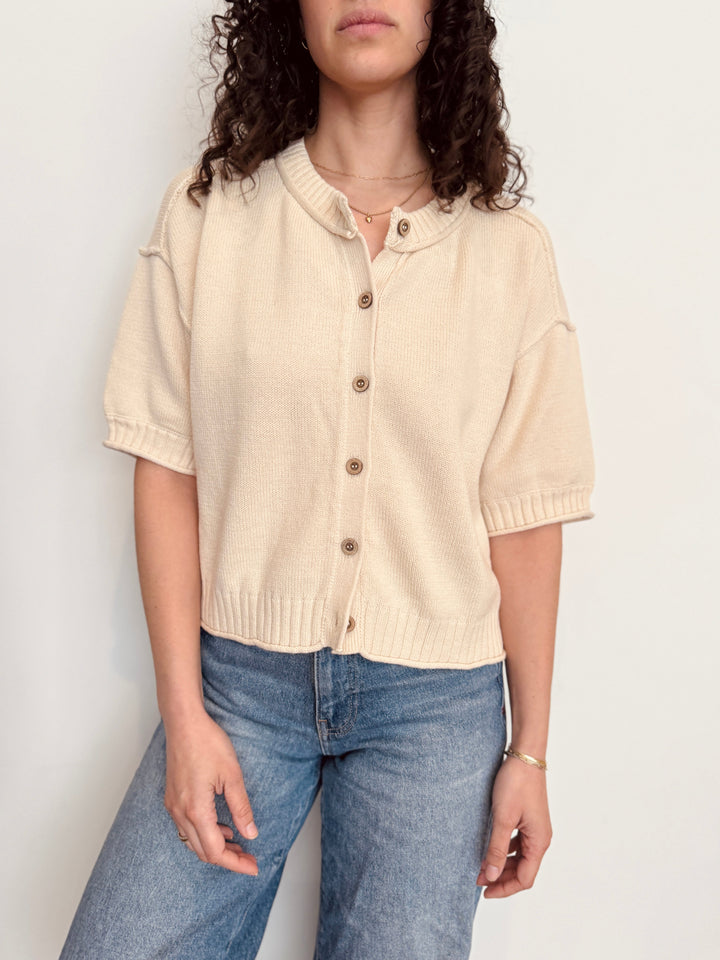 Person wearing a beige knitted cardigan and blue jeans against a plain background