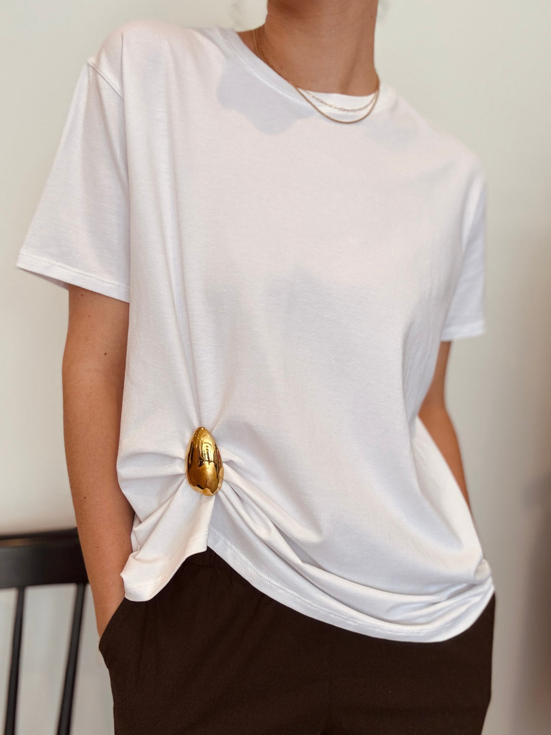 Person wearing a loose white tee with a gold drip shaped brooch pinning up the lower hem of the tee.