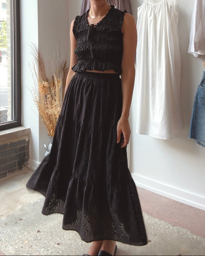 Woman wearing a black eyelet midi skirt standing in a clothing store with white walls and clothing racks.