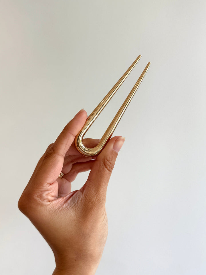 Hand holding a gold hair fork pin against a plain background