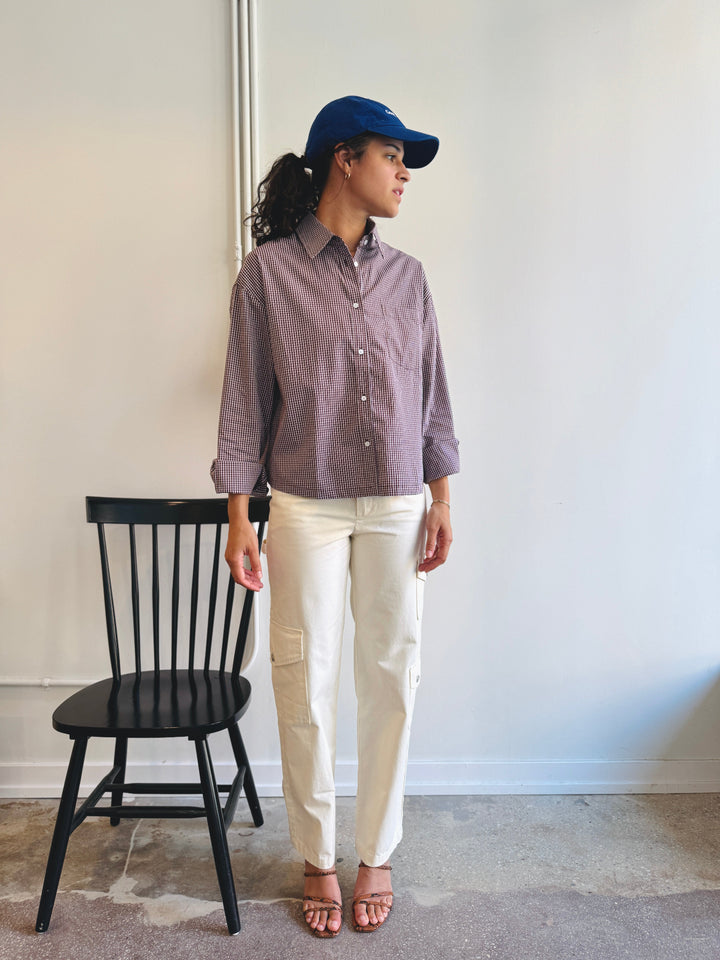 Person wearing a brown gingham shirt, white pants, and a blue cap standing next to a black chair against a white wall.