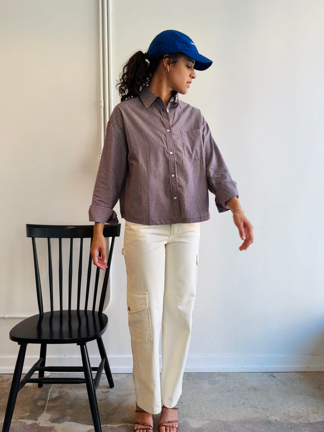 Person wearing a brown shirt and white pants standing next to a black chair against a white wall.