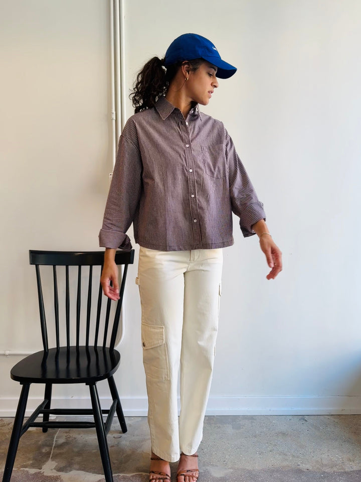 Person wearing a brown shirt and white pants standing next to a black chair against a white wall.