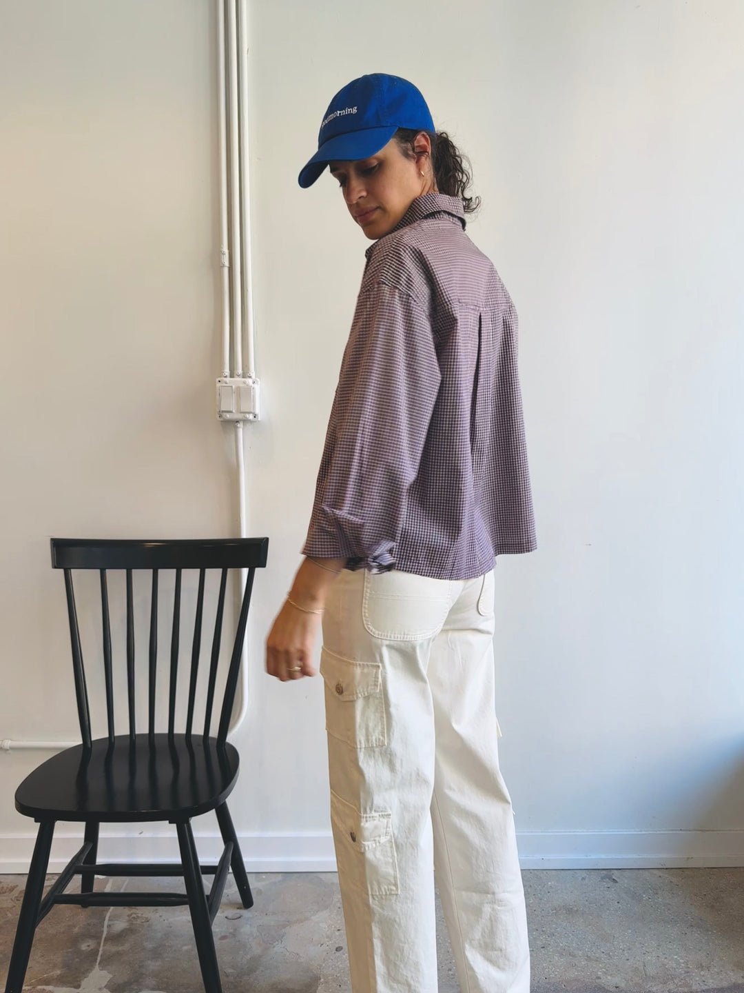 Person wearing a blue cap and brown gingham shirt standing next to a black chair against a white wall.