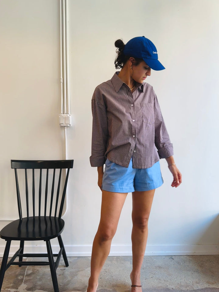 Woman wearing a brown button-up shirt, blue shorts, and a blue cap indoors.