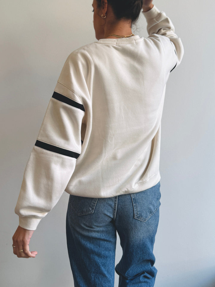 Person wearing a beige long-sleeve shirt with black stripes on sleeves and blue jeans against a plain background