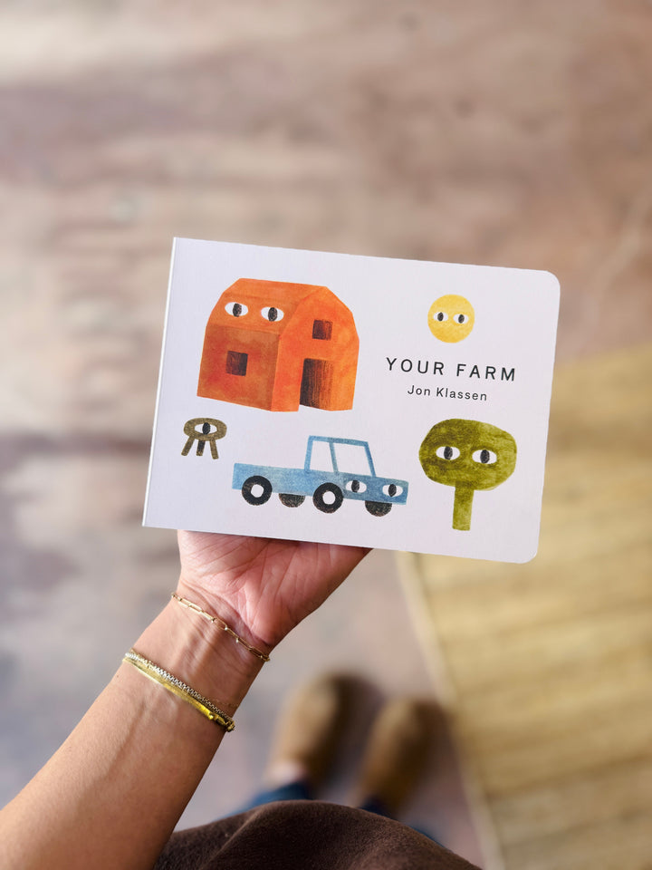 Hand holding a children's book titled 'Your Farm' by Jon Klassen with illustrations of a house, car, and tree.